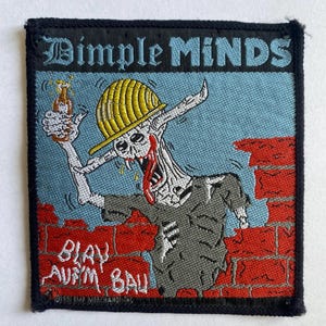 May include: Embroidered patch with the words "Dimple Minds" in gothic font. The patch features a cartoon zombie construction worker holding a beer bottle, with the text "Blau Auf'm Bau" below. The background is blue with a brick wall.