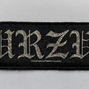 May include: Black rectangular patch with the word "BURZUM" in silver glitter gothic lettering. The patch has a black border and the text "©1997 MISANTHROPY RECORDS" is printed below the word.