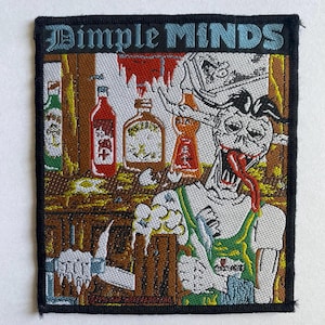Può includere: Toppa ricamata con la scritta "DIMPLE MINDS" in blu. L'immagine raffigura uno zombie dei cartoni animati con una lunga lingua rossa, bottiglie e un boccale di birra. La toppa ha un bordo nero.