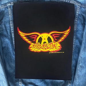 May include: A black patch with the Aerosmith logo in yellow, red, and orange, with the band's name in a stylized font. The patch is rectangular and is placed on a denim jacket.