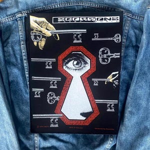 May include: A black graphic with the text "SCORPIONS CRAZY WORLD", a keyhole design, and keys. The design includes an eye within the keyhole and a hand holding a key. The graphic is on a denim jacket.
