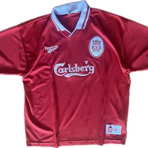 May include: Red Liverpool soccer jersey with white collar and sleeve trim. Features the Reebok logo, the Carlsberg logo, and a team crest. Includes a small tag with text.