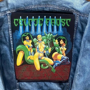 May include: A light blue denim jacket with a large patch on the back. The patch displays the text "CELTIC FROST" in green, above a graphic of three figures and a horned creature in a fantasy scene.