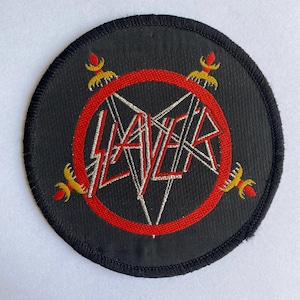Könnte beinhalten: Ein runder, schwarzer Aufnäher mit dem Wort "SLAYER" in einer stilisierten, eckigen Schriftart, mit einem roten Ring und weißen Linien. Das Design umfasst vier goldene und rote dekorative Elemente. Der Aufnäher hat einen schwarzen Rand.