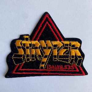 May include: Embroidered patch with the word "STRYPER" in yellow and black, set on a black triangle with red trim. The text "ISAIAH 53:5" is below the main text. The patch has a black border.
