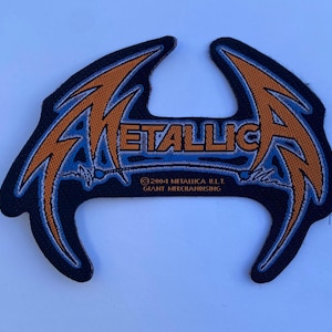 May include: A black embroidered patch featuring the Metallica logo in orange and blue. The text reads METALLICA with the copyright 2004 METALLICA U.I.T. GIANT MERCHANDISING printed below. The design has sharp, angular edges and a textured fabric finish.