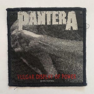 May include: A square fabric patch featuring a black and white graphic of a fist. The word PANTERA is printed in large white letters at the top. The text VULGAR DISPLAY OF POWER is printed in red at the bottom, with 1992 PANTERA text below it.