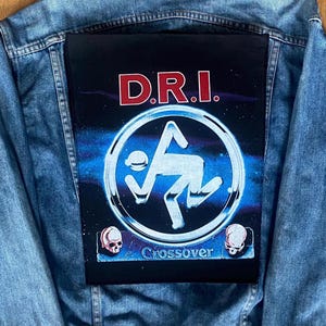 Peut inclure: Veste en jean bleu clair avec un grand écusson noir dans le dos. L'écusson présente le nom du groupe "D.R.I." en lettres rouges au-dessus d'un graphisme argenté et bleu. Le mot "Crossover" et deux illustrations de crânes sont en dessous.