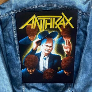 May include: Pale blue denim jacket with a large black patch. The patch displays the band name "ANTHRAX" in yellow. The image shows a man in a suit and hat surrounded by figures with brown hair.