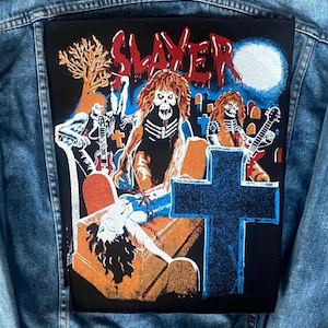 May include: A black patch with the word "SLAYER" in red, showing a graveyard scene with skeletons, a cross, and a full moon. The patch also depicts a person on a tomb. It is sewn onto a denim jacket.