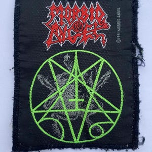 May include: A rectangular black fabric patch featuring the red Morbid Angel logo at the top with the text 1991 MORBID ANGEL printed vertically on the right side. Below the logo is a bright green embroidered pentagram design on a black background.