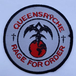 May include: A circular embroidered patch with a light gray background and a black stitched border. It features the red text "QUEENSRŸCHE" and "RAGE FOR ORDER" surrounding a central black winged emblem positioned above a red globe graphic.