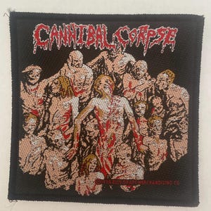 May include: Black square patch featuring the band name "CANNIBAL CORPSE" in red, stylized lettering. The patch depicts a group of figures with blood-like red accents. The patch is likely for a jacket or bag.