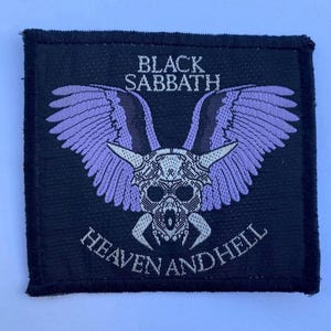 Könnte beinhalten: Ein quadratischer schwarzer Stoffaufnäher mit einer Stickerei eines geflügelten Schädels mit Hörnern. Der Text BLACK SABBATH ist in Weiß über dem Schädel gestickt, und HEAVEN AND HELL ist in Weiß unter dem Schädel in einem Bogen gestickt.