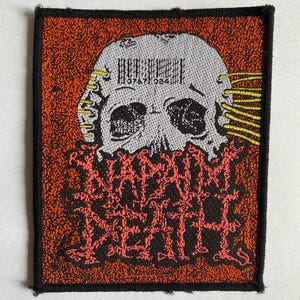 May include: A rectangular embroidered patch featuring a light gray skull on a textured orange background. The skull has yellow horizontal lines on the sides and a barcode above the eye sockets. Below the skull is the text NAPALM DEATH in red.