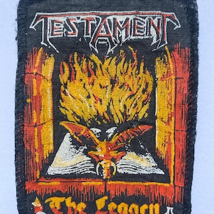 May include: Black patch with the band name "TESTAMENT" above a fiery image of a bat-like creature with a book and flames. The text "The Legacy" is below the image, with a skull and bottle graphic.