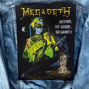 May include: A black patch with the Megadeth logo in yellow, and an illustration of a skeletal soldier in green and yellow. The text on the patch reads "SO FAR. SO GOOD.. SO WHAT!" The patch is attached to a denim jacket.