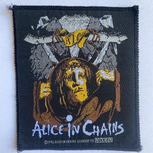 Pode incluir: Patch retangular preto com o nome da banda "Alice in Chains" em escrita branca. O patch apresenta uma imagem central de uma figura com rosto amarelo e cabelo castanho. A imagem está sobre um fundo texturizado.