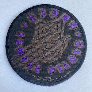 Pode incluir: Um patch circular preto com um personagem de desenho animado marrom com um chapéu e o texto roxo "STONE TEMPLE PILOTS". O patch também inclui o texto "©1993 STONE TEMPLE PILOTS THE GIANT MERCHANDISING COLLECTION."