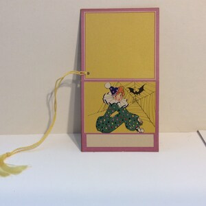 May include: A pink and yellow card with a clown in a green and white polka dot costume sitting in a spider web. A bat is flying above the clown. The card has a blank space for writing.