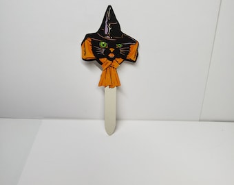 May include: Black cat wearing a witch's hat and orange scarf. The cat is a Halloween decoration made of cardboard.