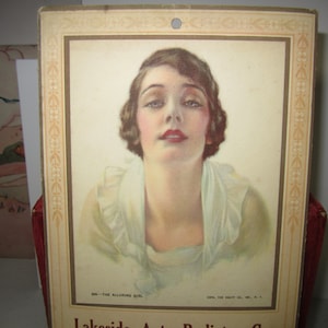 May include: Vintage print featuring a woman with dark hair, red lips, and a light-colored dress. The print is framed with a decorative border and includes the text "ON - THE ALLURING GIRL" and "Lakeside Auto Polishing Co."