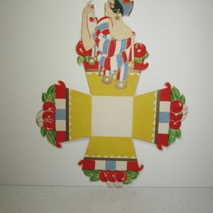May include: A vintage paper die-cut decoration featuring a woman in a patriotic outfit, holding cherries. The figure is atop a yellow box with red, white, and blue accents and cherry clusters. The design has a retro, art deco style.