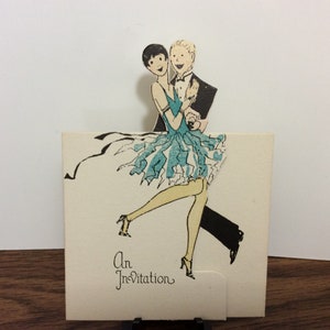 May include: A vintage invitation card with a pop-up illustration of a couple dancing. The woman is wearing a blue and white dress and the man is wearing a black suit. The text "An Invitation" is printed at the bottom of the card.