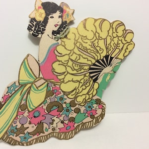 May include: A vintage paper doll cutout of a woman wearing a pink and yellow dress with a large yellow fan. The doll has black hair and a gold crown.