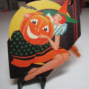 May include: Vintage Halloween die-cut decoration featuring a smiling jack-o'-lantern and a witch in an orange dress and green hat. The background includes a yellow moon and a black bat. The decoration is on a black tag with orange accents.