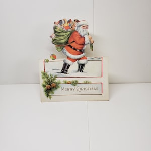 Vintage 1920's-30's unused colorful die cut Christmas place card Santa Claus walking in snow with his sack of toys, ball and book fall out