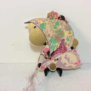 May include: A vintage paper ornament featuring a whimsical design of a pink and purple seashell with a gold and green sail. The ornament is suspended from a pink tassel.