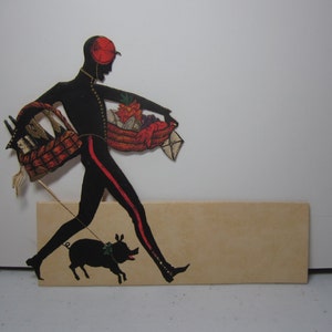 May include: A black silhouette of a person wearing a red hat and carrying a basket of fruit and vegetables. The person is walking a black pig on a leash. The silhouette is on a beige background.