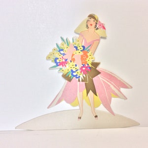 May include: A cutout of a woman in a pink dress and a yellow hat holding a bouquet of colorful flowers. The woman is standing on a white platform.