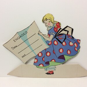 May include: A vintage cardstock cutout of a girl in a blue polka dot dress holding a card that says "ENGAGED" with lines for the date and names.