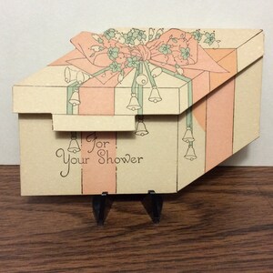 May include: A light pink and white gift box with a green bow and bells. The box is open and the text "For Your Shower" is visible.