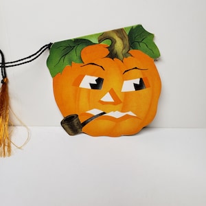 May include: A vintage Halloween decoration featuring a cartoon pumpkin with a grumpy expression, wearing a pipe. The pumpkin is orange with green leaves and a brown stem. The decoration is made of paper and has a string attached for hanging.