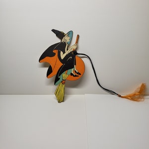 May include: A vintage Halloween decoration featuring a witch on a broomstick. The witch is wearing a black dress and hat, and has a long, flowing orange cape. The broomstick is yellow and has a green handle. The decoration is made of cardboard and has a string attached to it.