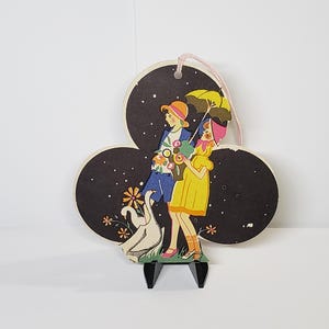 May include: Vintage decorative wooden cutout featuring a couple under a starry night sky. The couple is dressed in colorful attire, with one holding flowers and the other an umbrella. The cutout is mounted on a black stand and has a pink string for hanging.