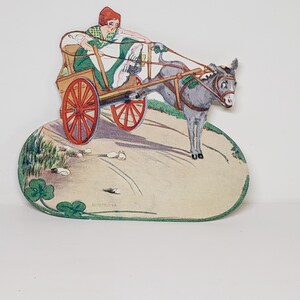 May include: A vintage paper cutout of a woman in a green dress and red hat riding a donkey pulling a cart. The donkey is gray and white, and the cart has red wheels. The cutout is on a tan background with green shamrocks.