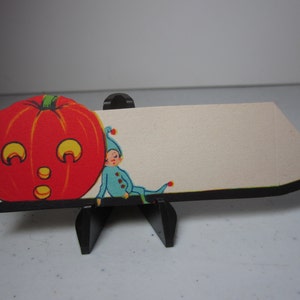 May include: A vintage Halloween-themed card featuring a smiling orange pumpkin with yellow accents and a small elf in blue attire. The card is mounted on a black stand, perfect for seasonal decor.
