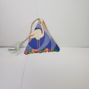 May include: A triangular-shaped paper ornament featuring a white stork with orange beak and legs, standing on a bed of flowers. The background is blue with a geometric pattern. A string and tassel are attached to the top.