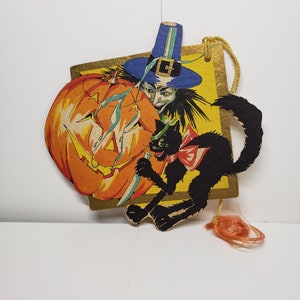 May include: A vintage Halloween card featuring a witch with a blue hat, a black cat, and a jack-o'-lantern. The card is yellow with gold accents and has a string attached for hanging.