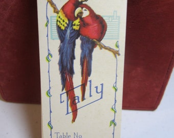 May include: A vintage white card with a blue border and a green vine design. The card features a drawing of two red and yellow parrots perched on a branch. The text "Tally" is written in blue on the card. The card also has the text "Table No." and "Couple No."