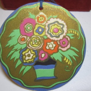May include: A round, blue and gold paper ornament with a floral design. The flowers are pink, yellow, orange, and white with green leaves. The flowers are in a blue and green pot.