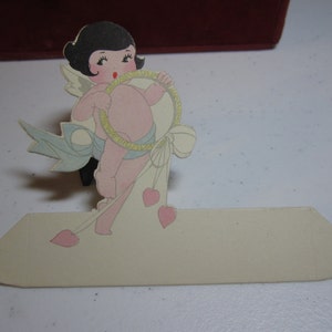 May include: A vintage Valentine's Day card featuring a cartoon cherub with black hair, holding a heart-shaped ring and wearing a blue ribbon. The cherub is standing on a white card with a blank space for writing a message.