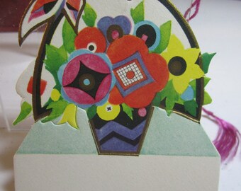 May include: A vintage paper gift tag with a colorful floral design. The tag features a bouquet of flowers in a vase with a geometric pattern. The tag has a hole at the top for string or ribbon.