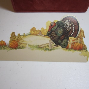 May include: A vintage Thanksgiving place card featuring a colorful illustration of a turkey standing on a fence with pumpkins and a farmhouse in the background.