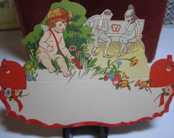 May include: A vintage Valentine's Day card with a red heart design. The card features a cupid with wings and a bow and arrow, a couple sitting on a bench, and a blank space for a message.