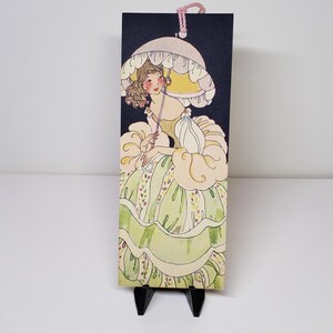 May include: A vintage-style bookmark featuring a woman with an umbrella. The illustration has a woman in a yellow dress and a matching umbrella. The bookmark is rectangular with a dark background and a pink string at the top.